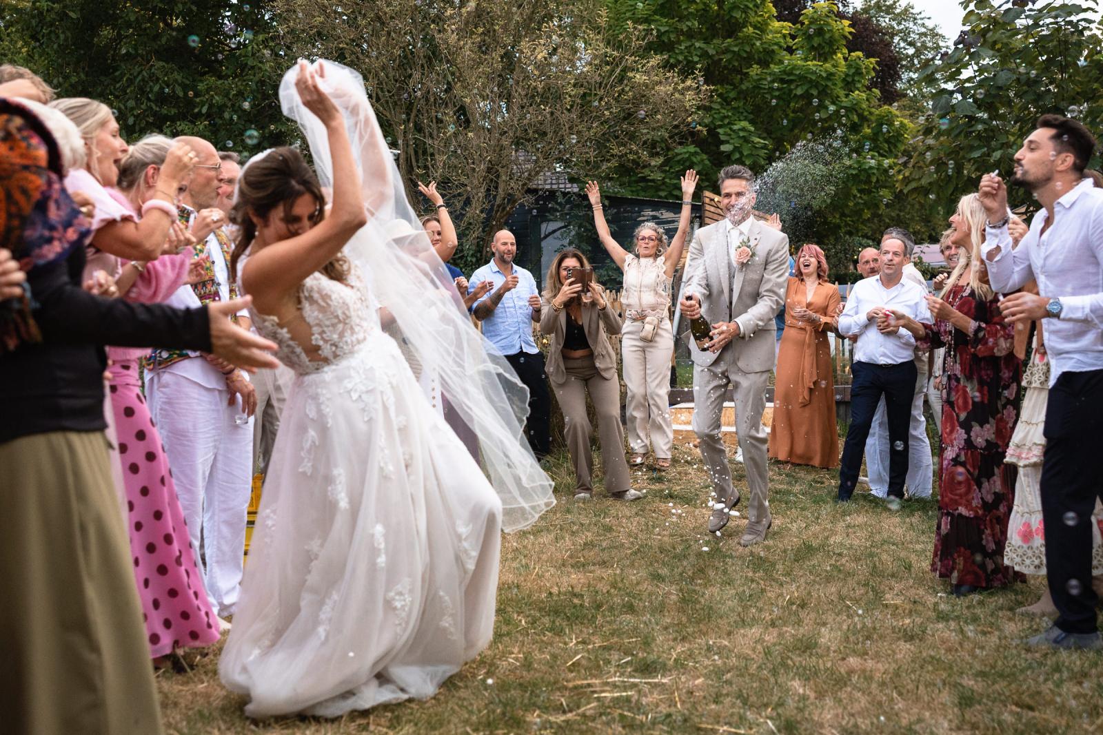 De bruid gooit haar sluier omhoog terwijl de bruidegom een fles champagne opent, omringd door feestende gasten.