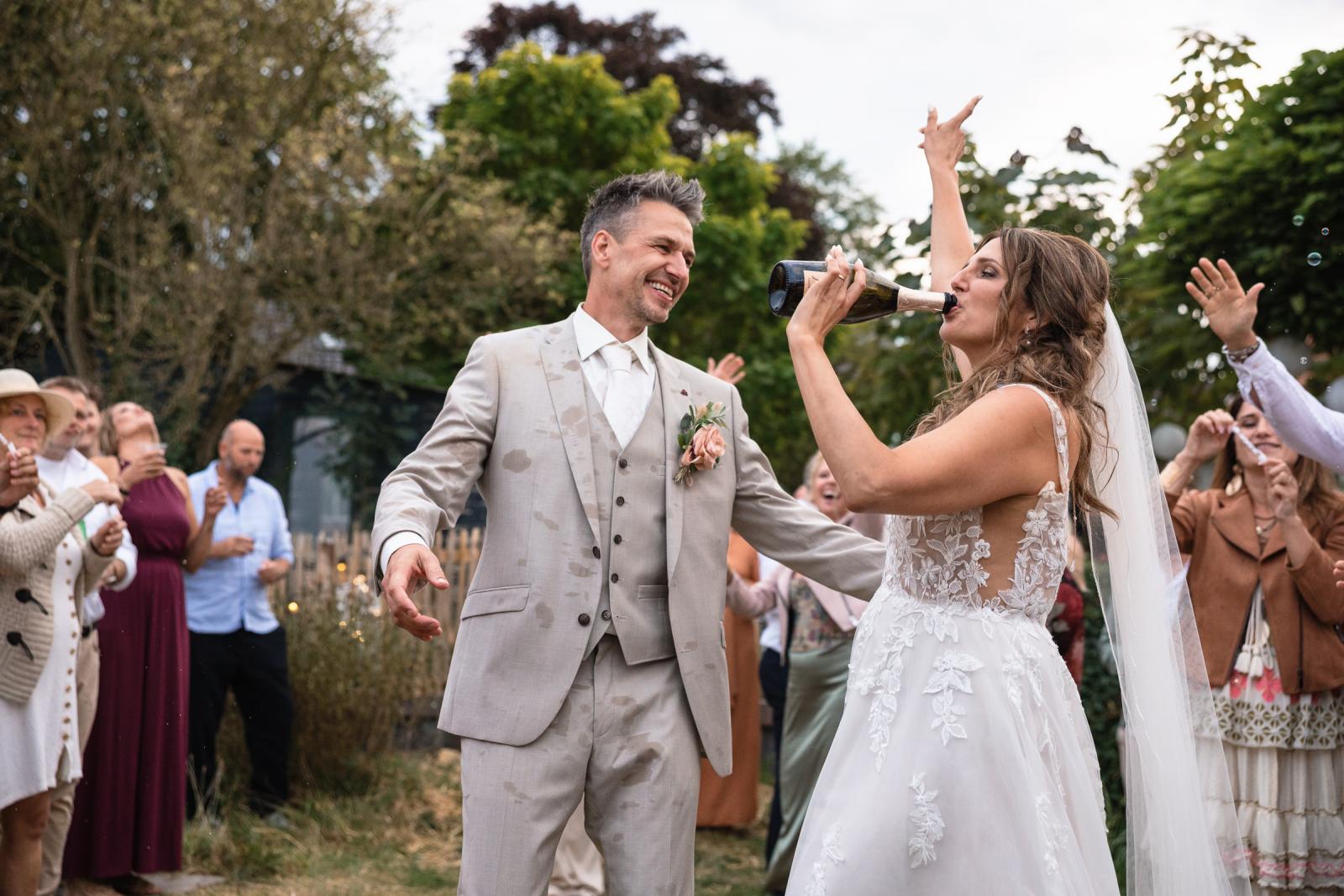 De bruid drinkt uit een champagnefles terwijl de bruidegom lachend toekijkt, midden tussen de gasten.
