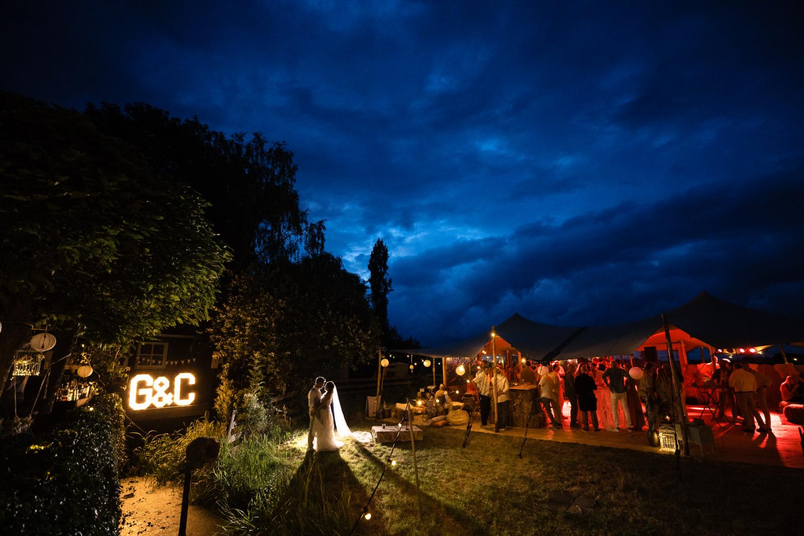 Avondshot met bruidspaar met de feesttent vol dansende gasten en een bruisende sfeer op de achtergrond.