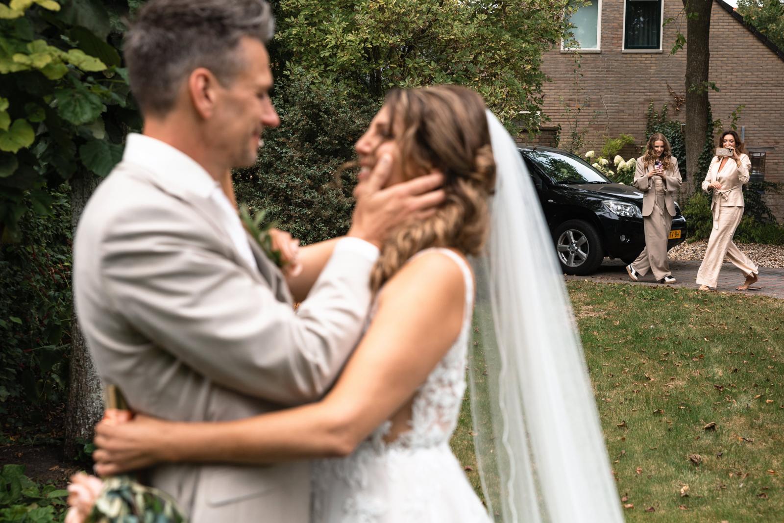 Gerrit en Chantal omhelzen elkaar liefdevol tijdens de first look, bekeken door vriendin en dochter op de achtergrond.