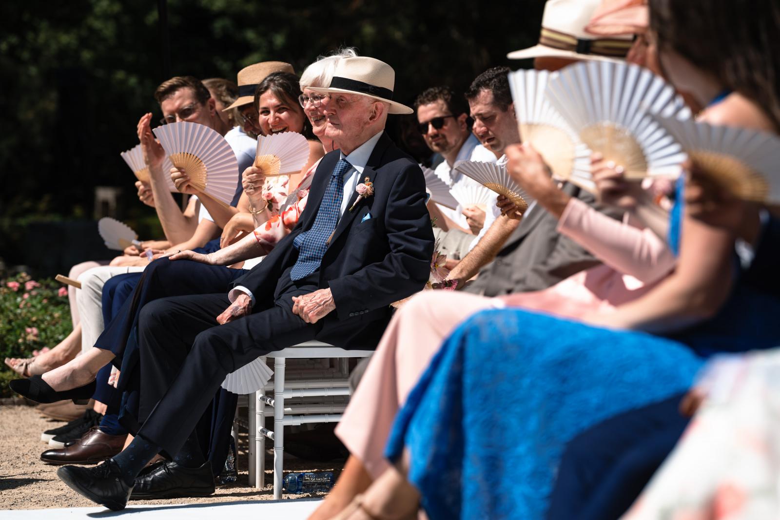 Oudere gast met hoed en strak pak geniet van de buitenceremonie in de zon.