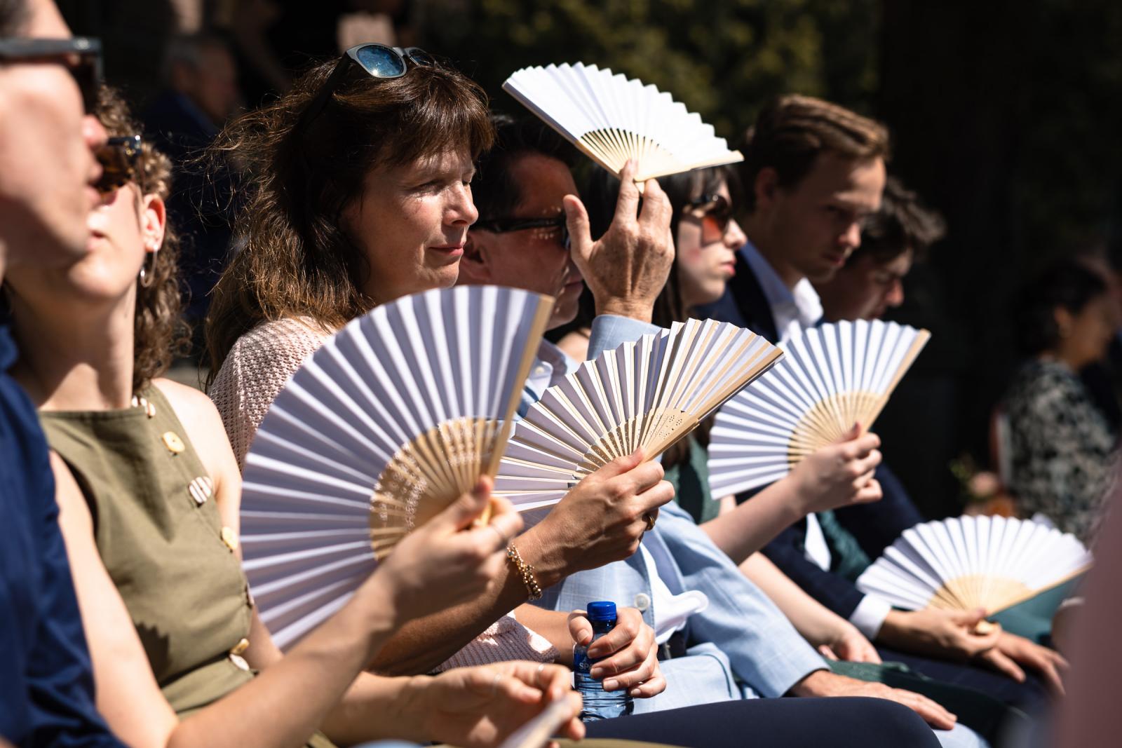 Huwelijksgasten zoeken verkoeling met witte waaiers tijdens een warme ceremonie op het landgoed.