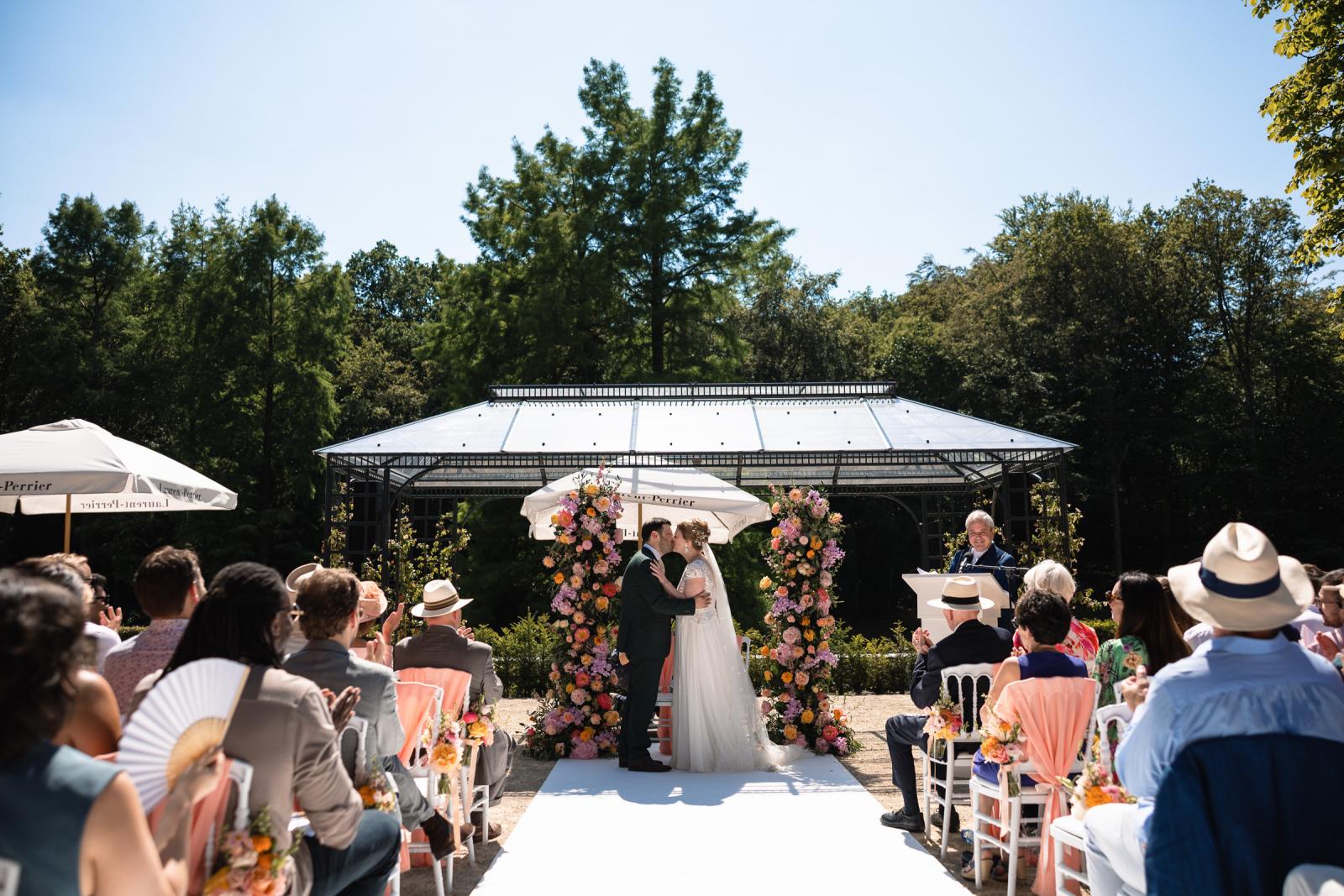 De eerste kus van bruid Hanneke en bruidegom Dennis onder de chuppah met bloemen.