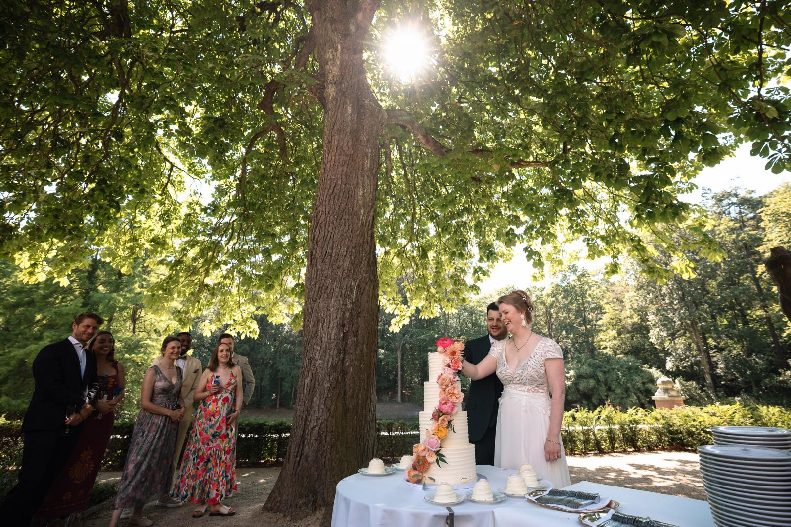 Aansnijden van de elegante trouwtaart met verse bloemen onder het bladerdak van een grote boom.