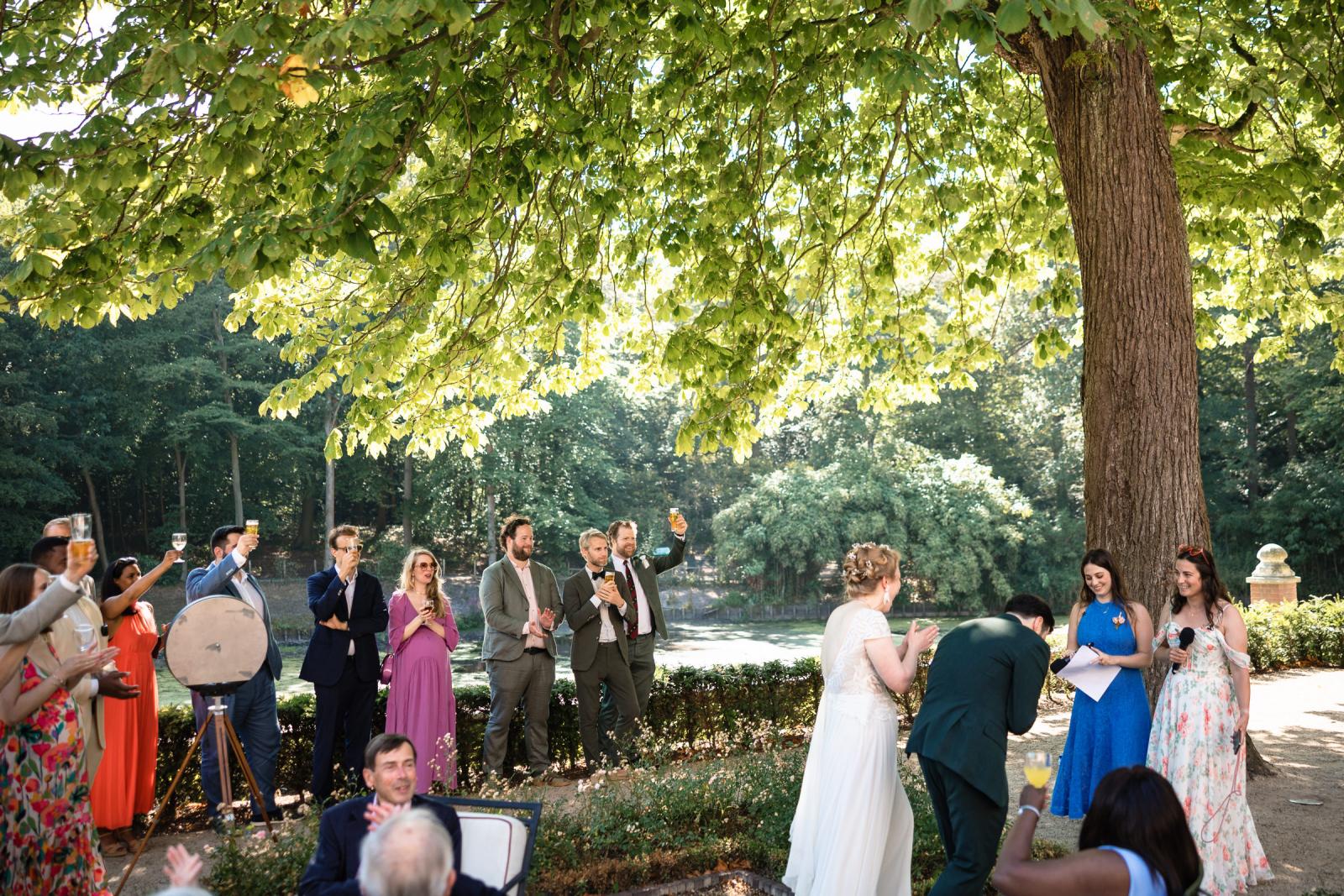 Proostmoment waarbij gasten het glas heffen op het bruidspaar tijdens een toespraak in de kasteeltuin.