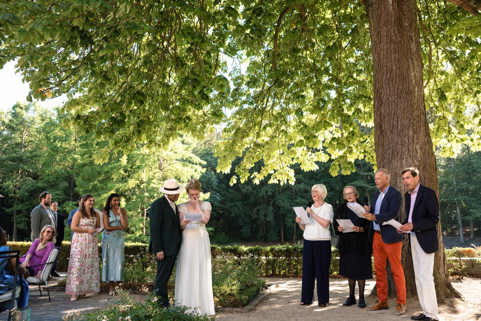 Gasten en familie dragen een speech en zingen voor bruidspaar onder een grote boom in de tuin van het landgoed.