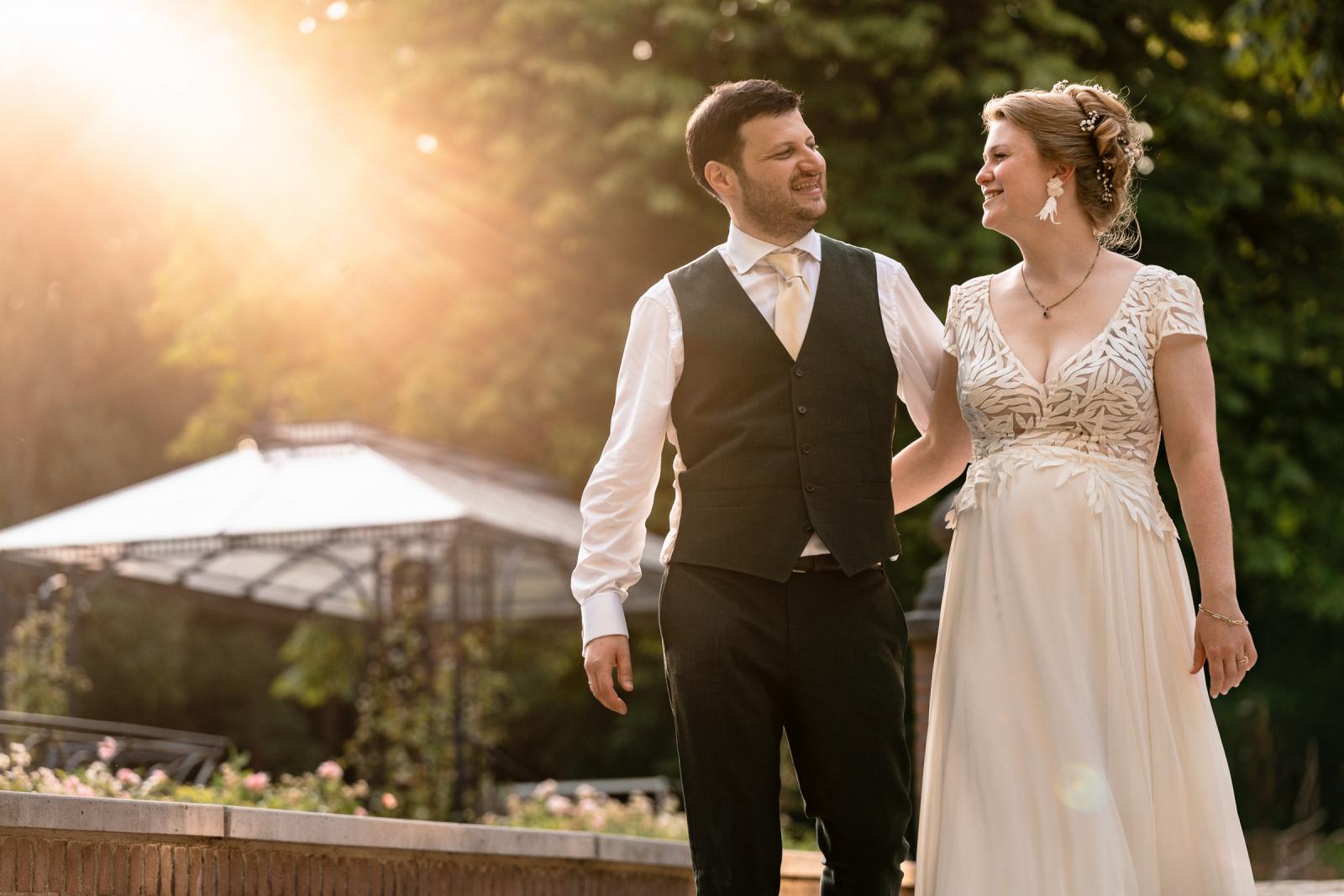 Lachend bruidspaar kijkt elkaar liefdevol aan tijdens een wandeling door de kasteeltuin tijdens de golden hour foto's