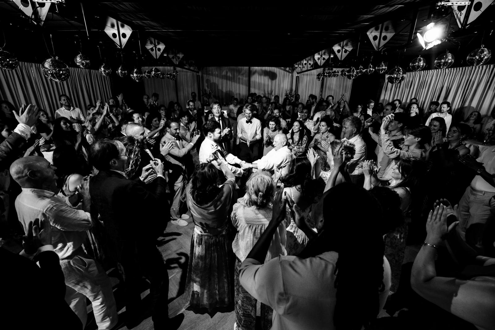 Zwart-witfoto van bruiloftsgasten die klappend in een cirkel om de dansende joodse mannen heen staan op de feestlocatie.