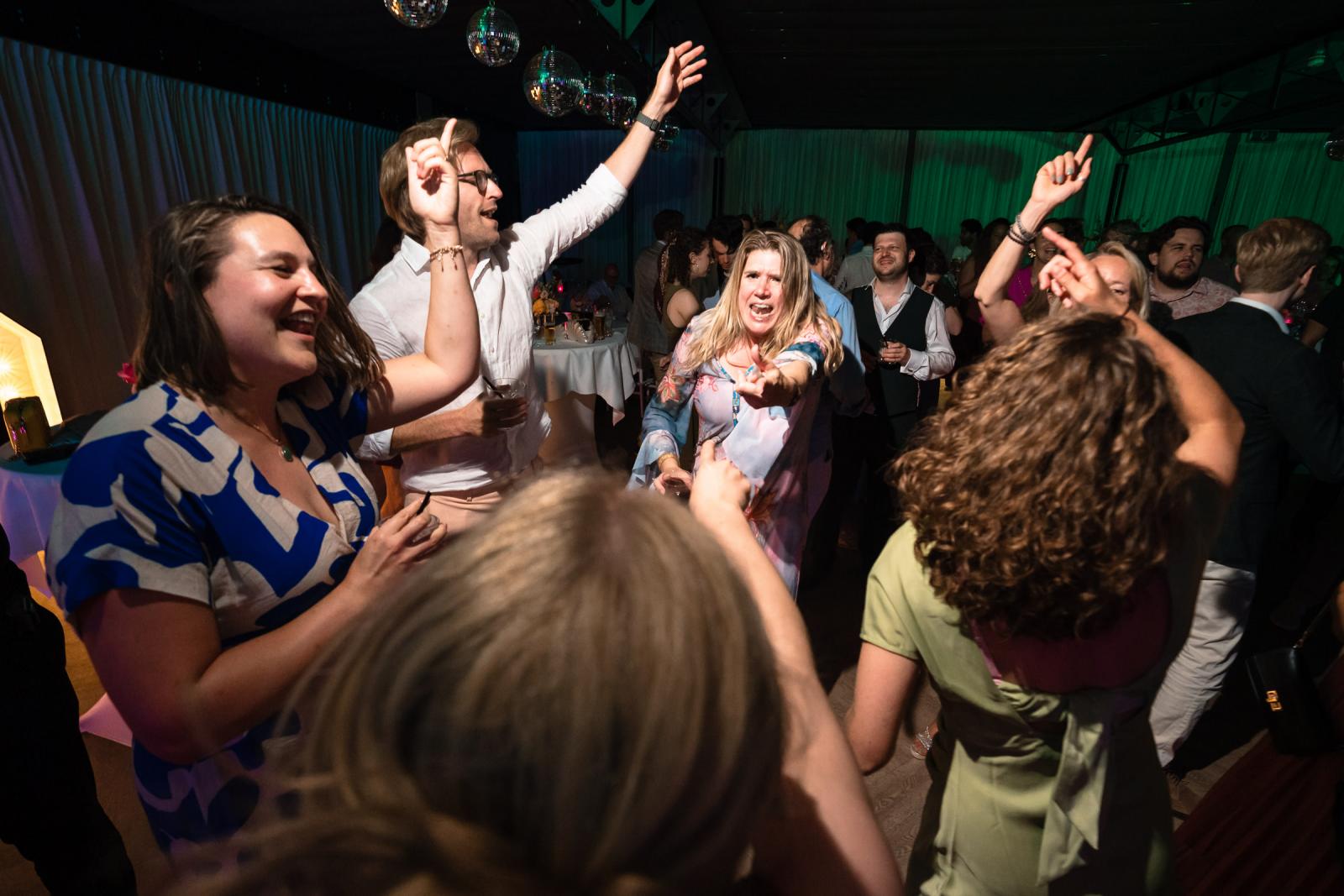 Vrolijke bruiloftsgasten dansen enthousiast met de handen in de lucht op de feestlocatie.