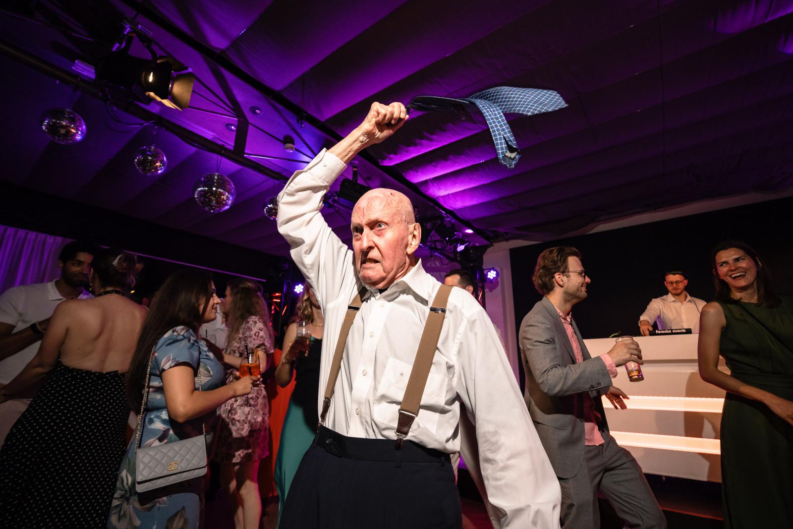 Oudere bruiloftsgast zwaait enthousiast met een stropdas boven zijn hoofd op het bruiloftsfeest