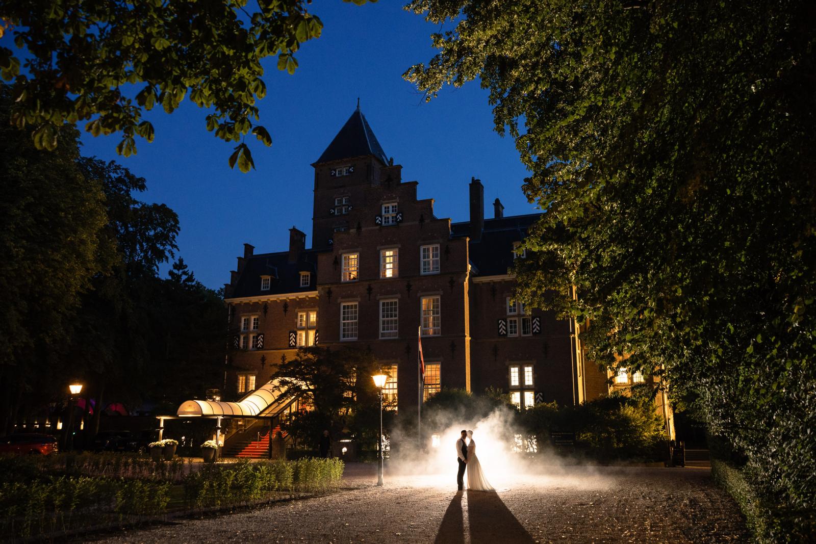 Magisch trouwportret van bruid en bruidegom in een mistwolk met tegenlicht voor het verlichte Kasteel