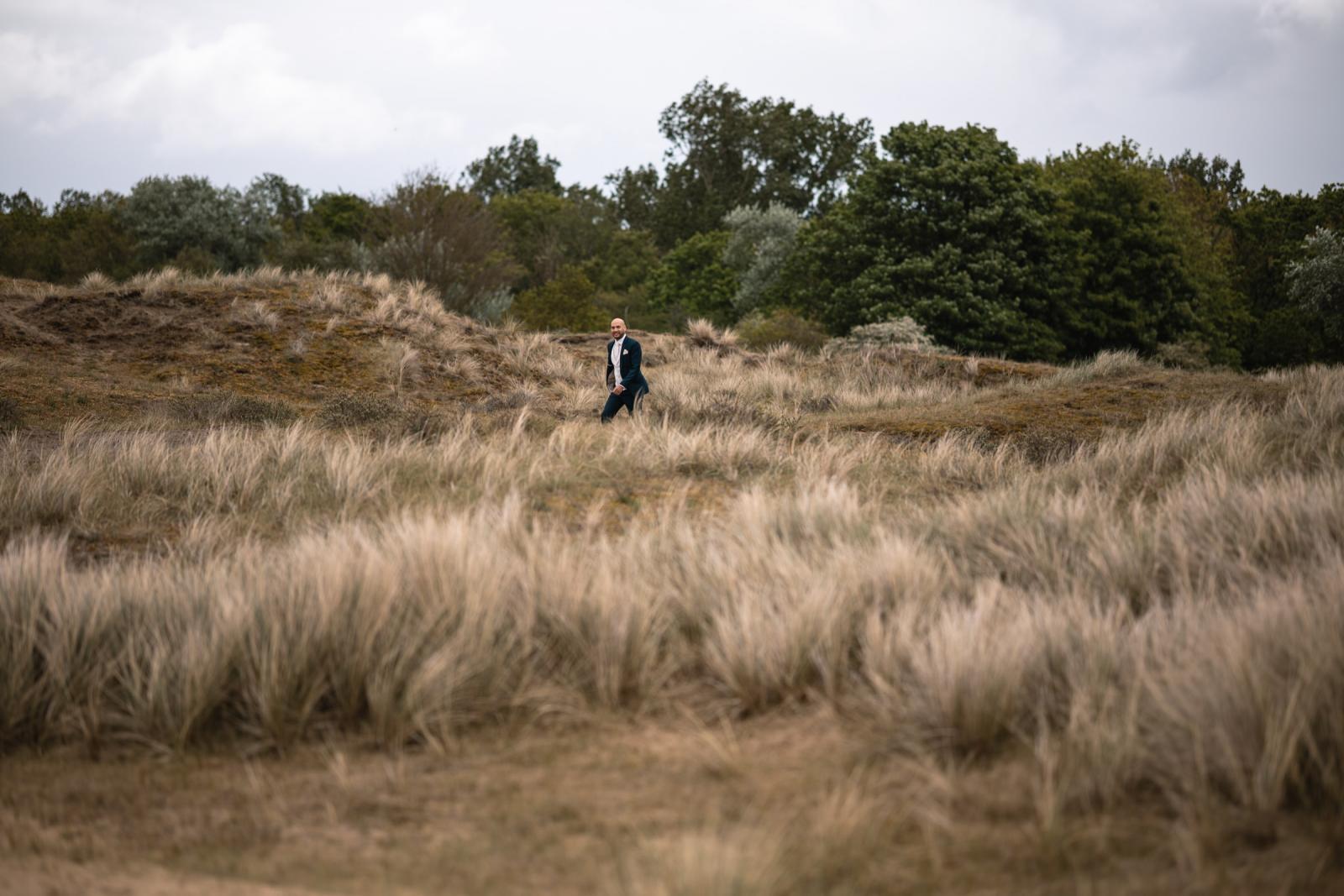 Bruidegom Mark loopt glimlachend in een donkerblauw trouwpak door het helmgras tijdens de first look in de duinen.