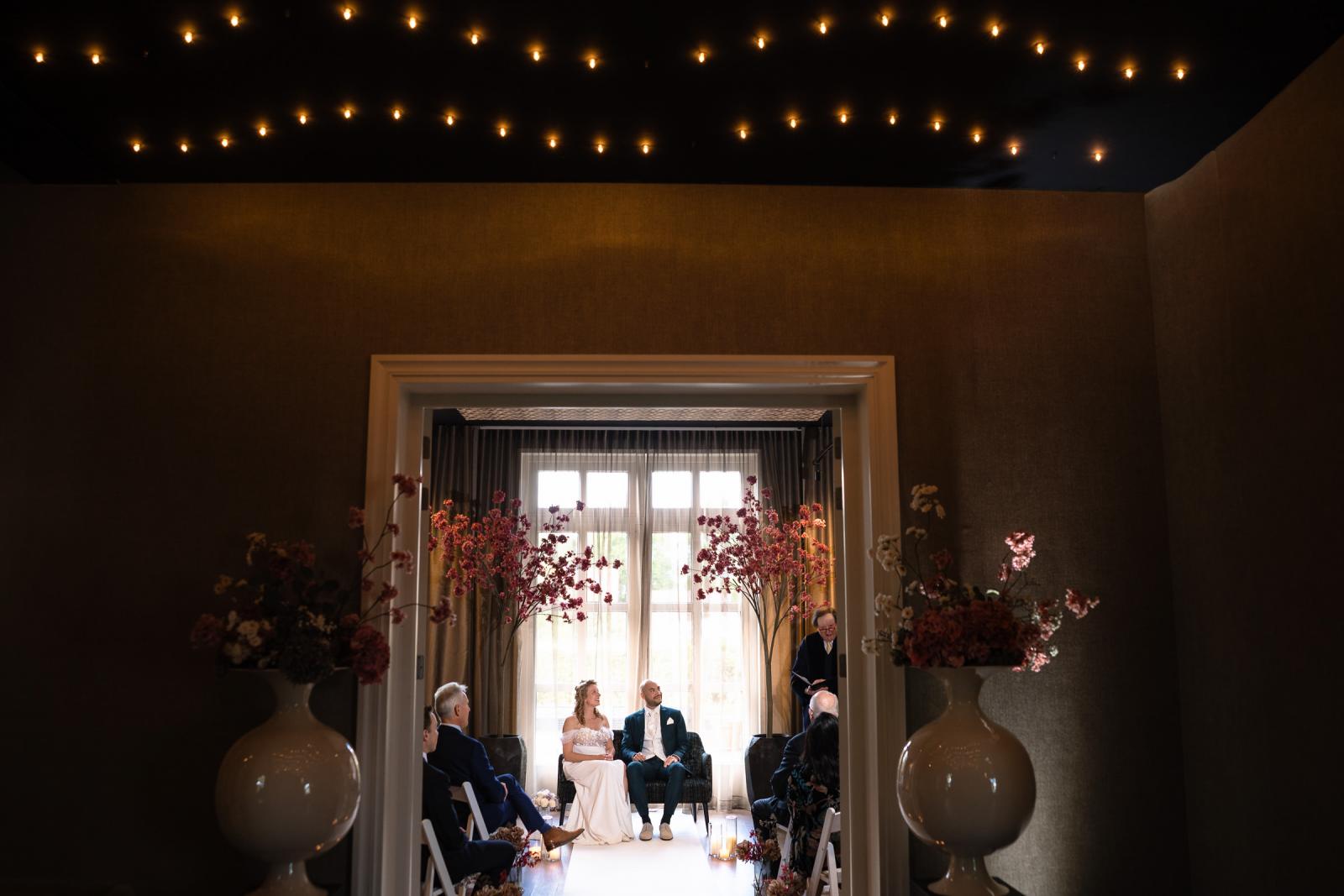 Een weidse overzichtsfoto van de intieme huwelijksceremonie in de warm ingerichte zaal met een indrukwekkende sterrenhemel van lichtjes op het plafond.