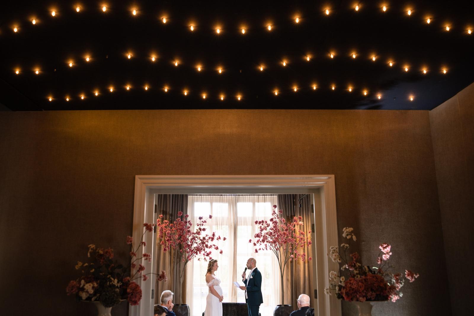 Een weidse foto van het bruidspaar en hun daggasten tijdens de geloften, omringd door de prachtige lichtjeshemel en de ceremoniedecoratie.