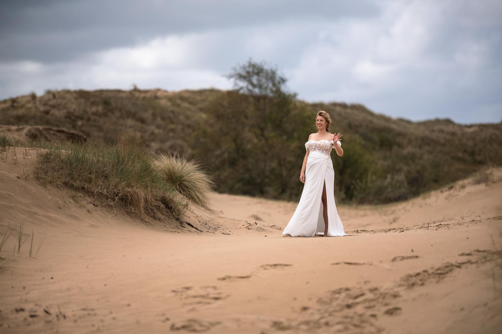 Bruid Judith zwaait enthousiast in haar trouwjurk op een zandpad in de Amsterdamse Waterleidingduinen.