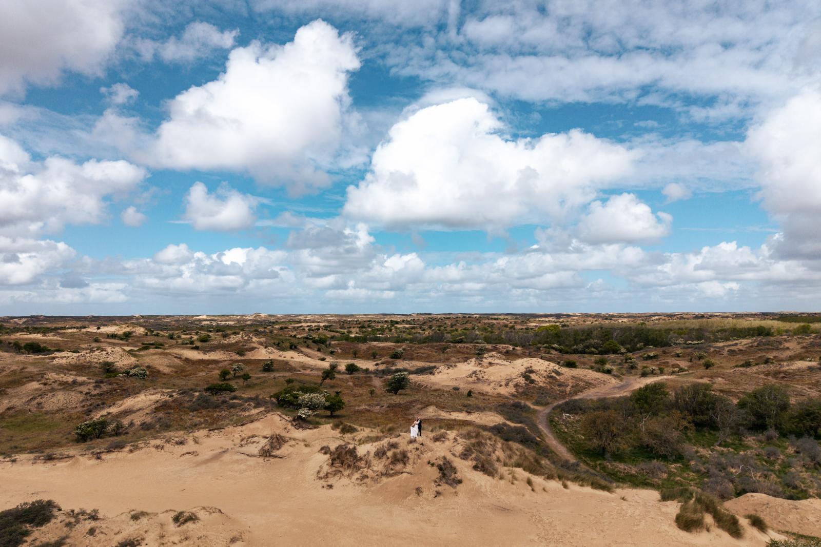 Prachtig weids overzichtsschot van het bruidspaar bovenop een duinpan onder een indrukwekkende wolkenlucht.