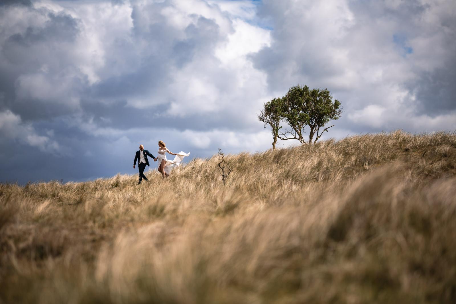 Bruidspaar Judith en Mark lopen hand in hand over de duinen onder een dramatische, bewolkte lucht.
