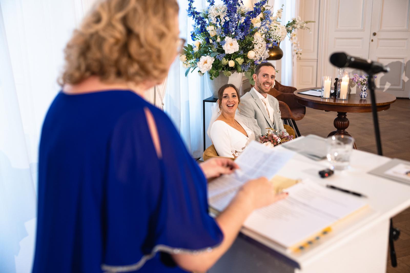 Bruidspaar lacht naar spreker tijdens ceremonie – persoonlijke trouwceremonie Spaansche Hof Den Haag.