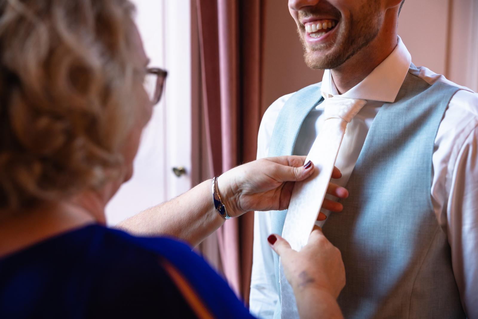 Close-up van moeder die de das van de bruidegom strikt – intiem voorbereidingsmoment Spaansche Hof Den Haag.