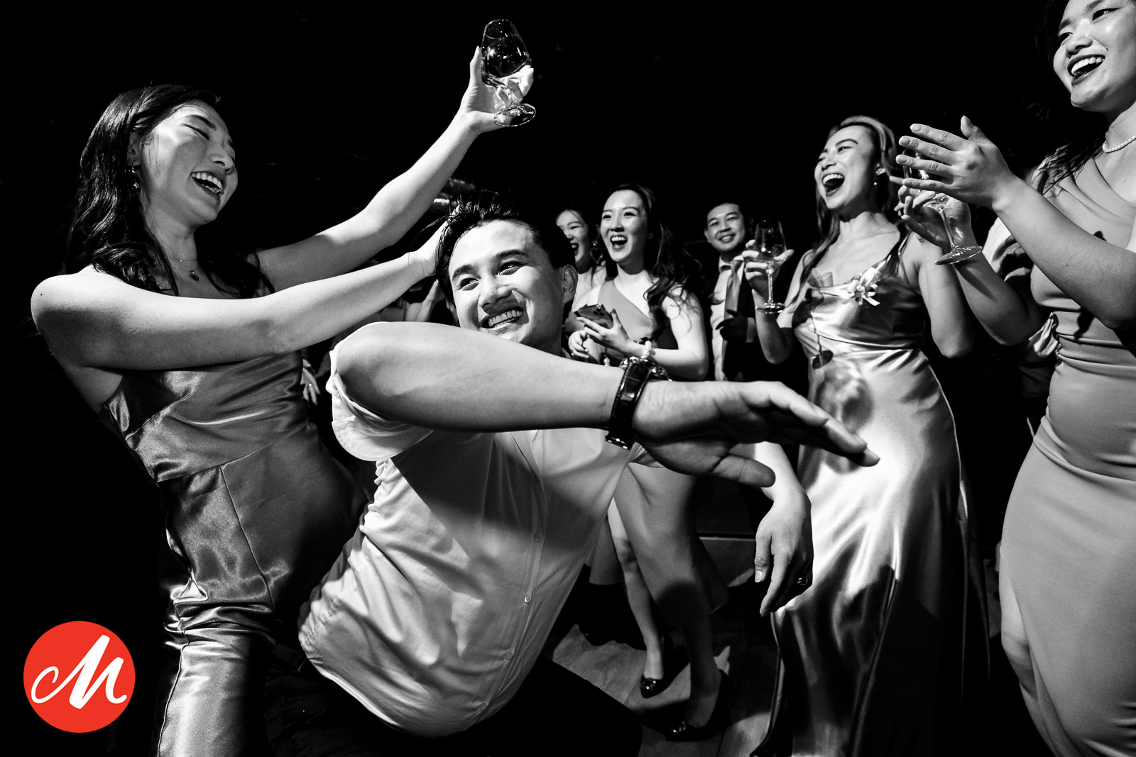 Zwart-wit trouwfotografie van een lachende bruidegom en dansende bruidsmeisjes tijdens een spetterend trouwfeest.