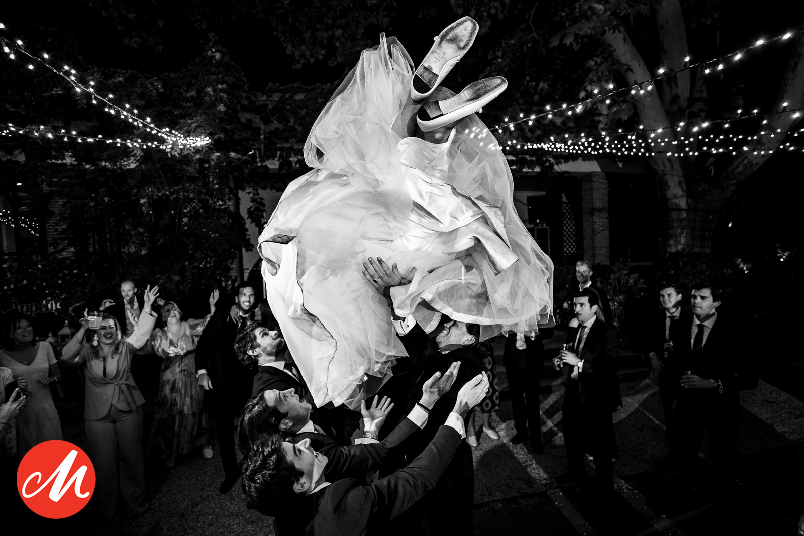 Spectaculaire zwart-wit actiefoto waarbij de bruid door bruiloftsgasten in de lucht wordt gegooid tijdens een buitenfeest.
