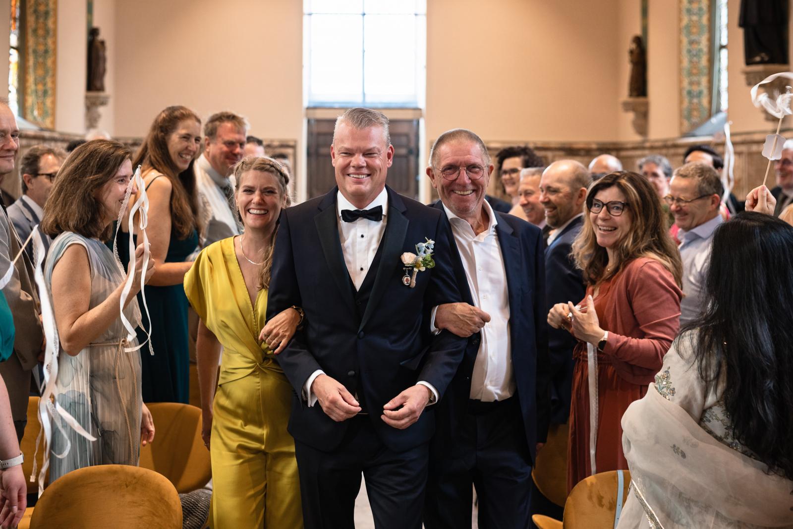Bruidegom Mike loopt arm in arm met dierbaren door de kapel voor de ceremonie