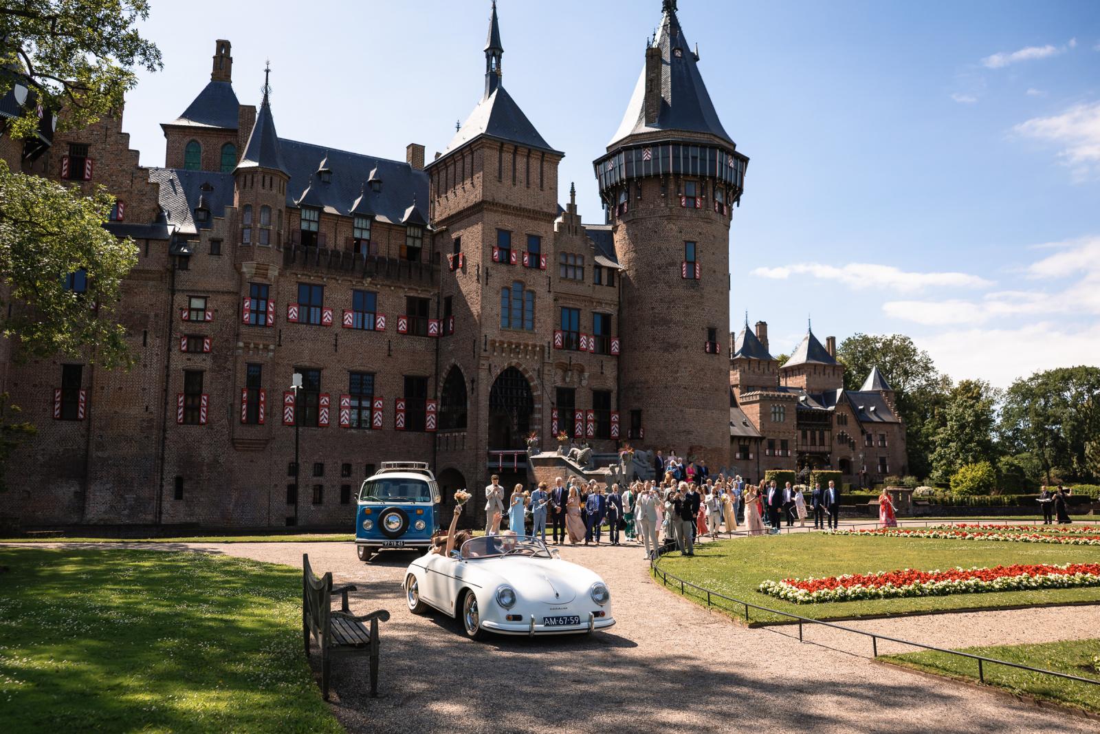 Bruidspaar rijdt in een klassieke witte Porsche cabrio voorbij het imposante Kasteel de Haar, gevolgd door een blauwe Volkswagen Bus.