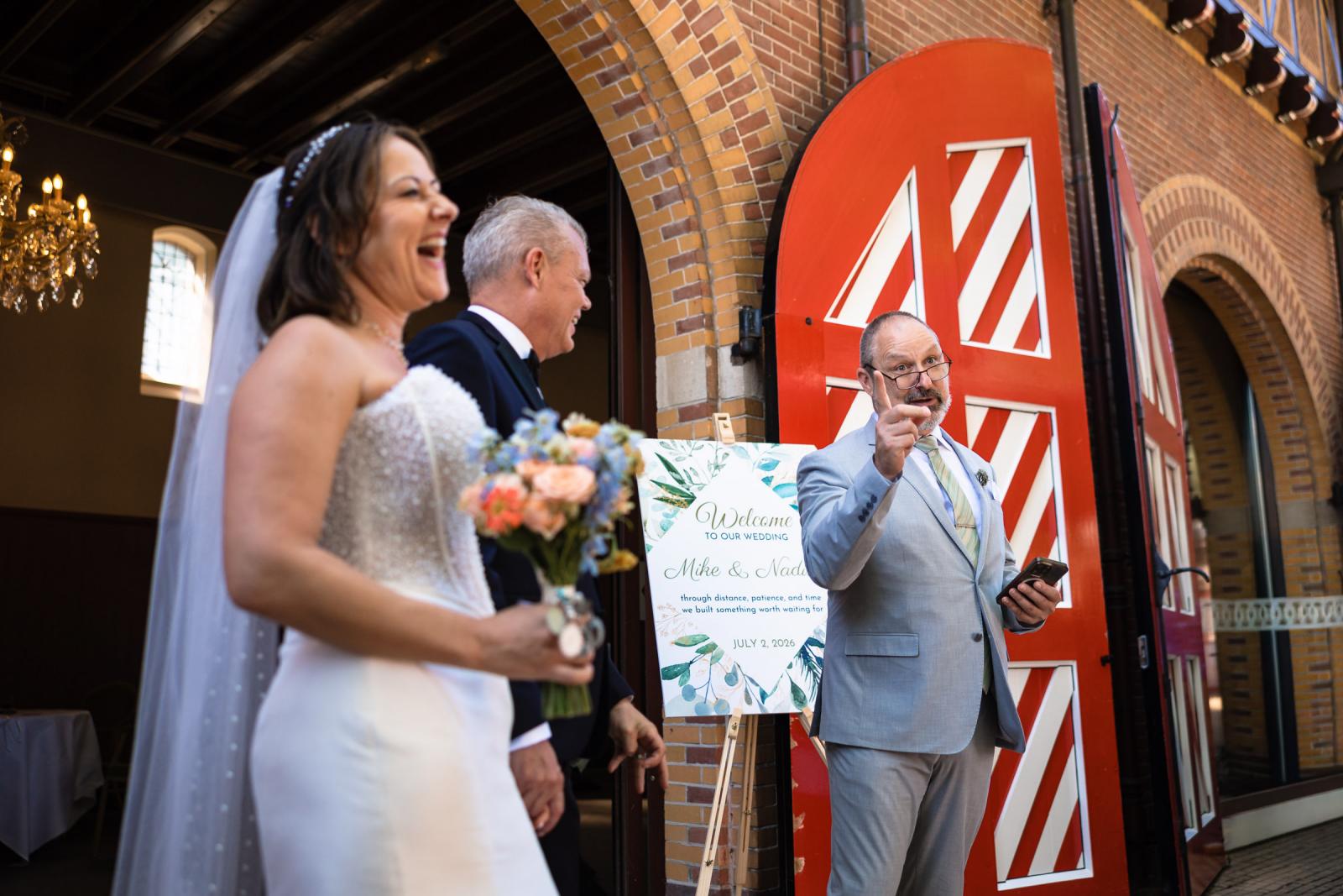 Lachende bruid Nadine met boeket en bruidegom Mike luisteren naar een speech bij de rode deuren van het Koetshuis van Kasteel de Haar.