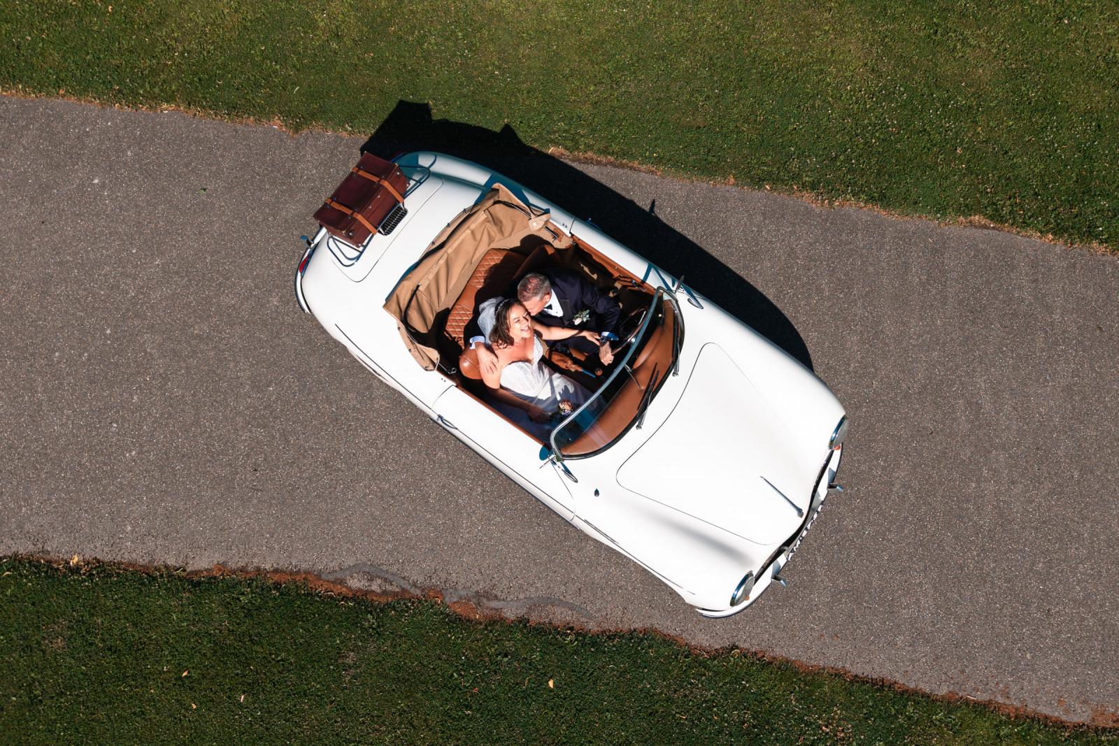 Luchtfoto van bovenaf van bruidspaar Mike en Nadine lachend in hun witte cabrio op het kasteelterrein.