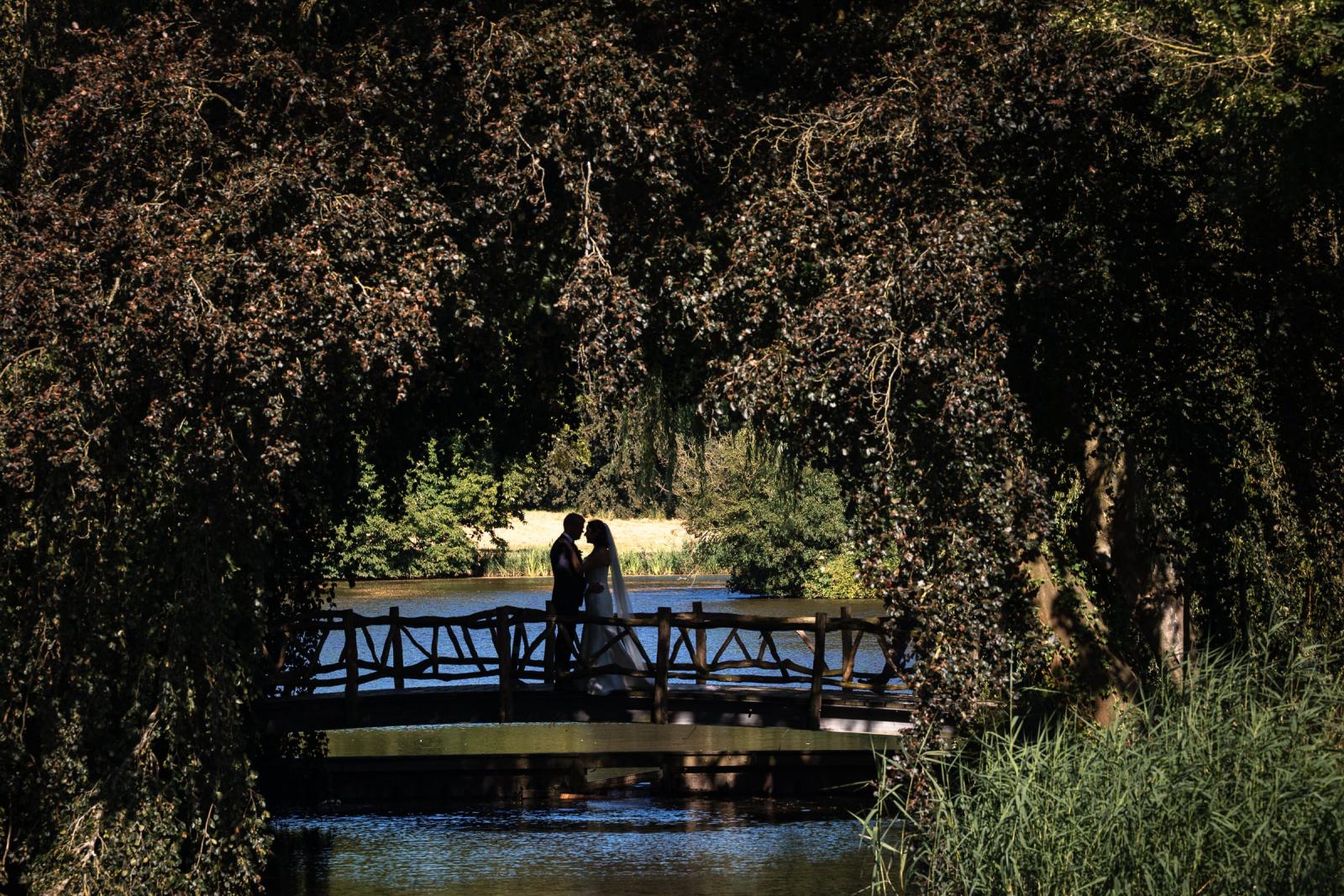 Sfeervolle silhouetfoto van het bruidspaar op een houten brug omringd door bomen bij het water van Kasteel de Haar.