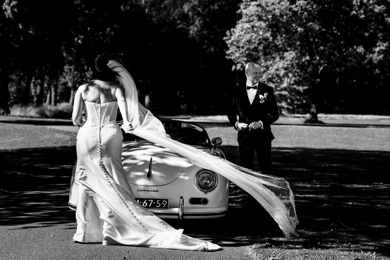 Zwart-wit foto van bruid Nadine met een lange wapperende sluier wandelend naar bruidegom Mike bij de Porsche.