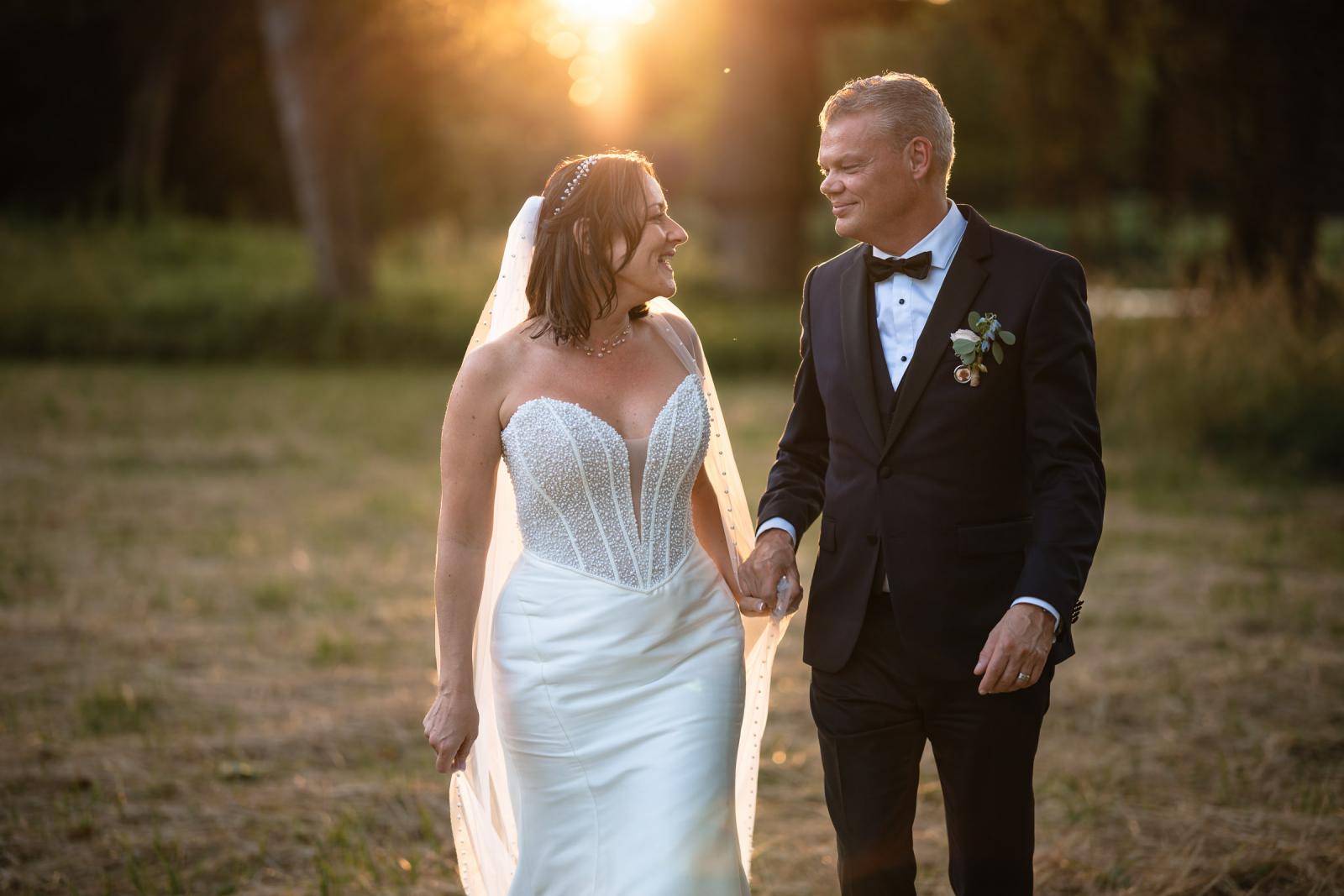 Bruidspaar Mike en Nadine lopen romantisch hand in hand door het park tijdens zonsondergang.