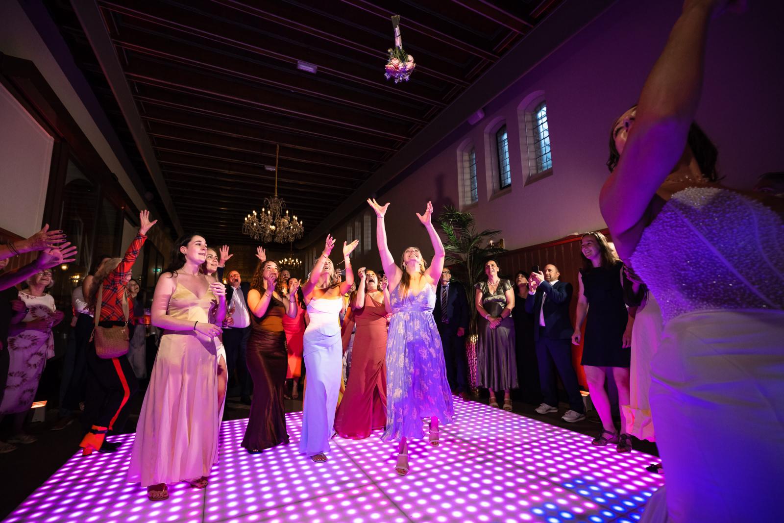 Actiefoto van het moment dat bruid Nadine haar trouwboeket in de lucht gooit naar een groep enthousiaste dames.