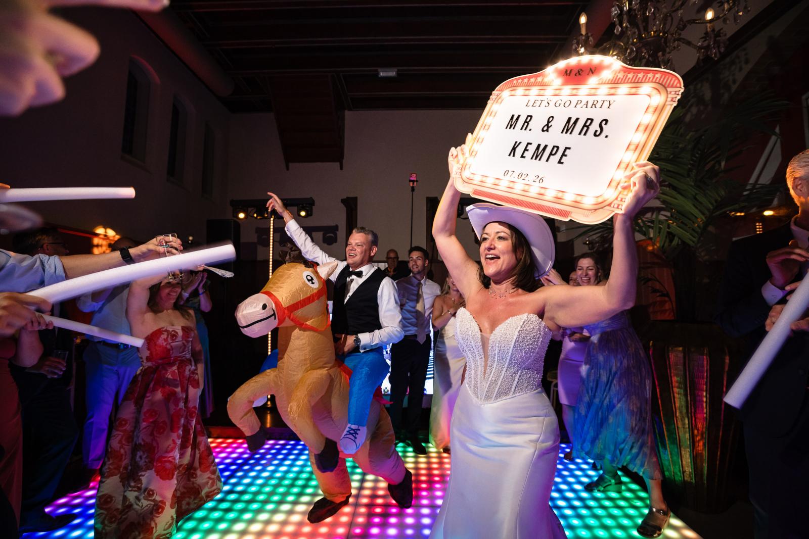 Bruid Nadine houdt een verlicht bord omhoog met 'MR. & MRS. KEMPE' terwijl bruidegom Mike op een opblaasbaar paard over de dansvloer feest.