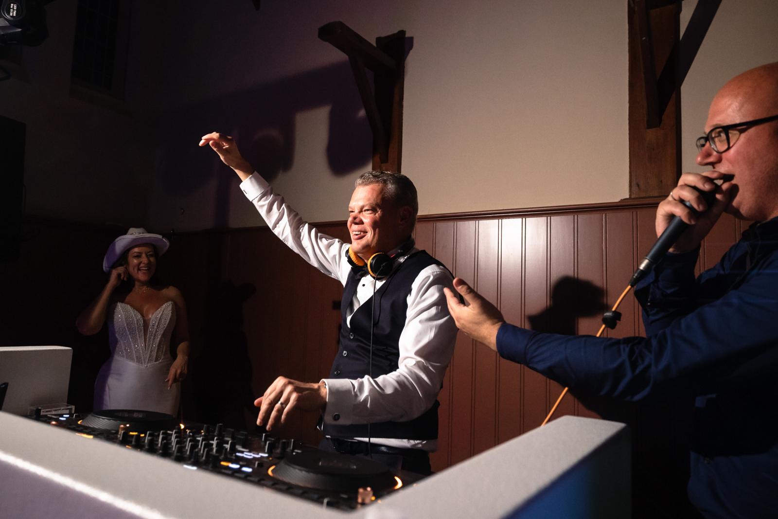 Bruidegom Mike neemt met een grote glimlach de DJ-booth over met een koptelefoon om zijn nek, gadegeslagen door bruid Nadine.