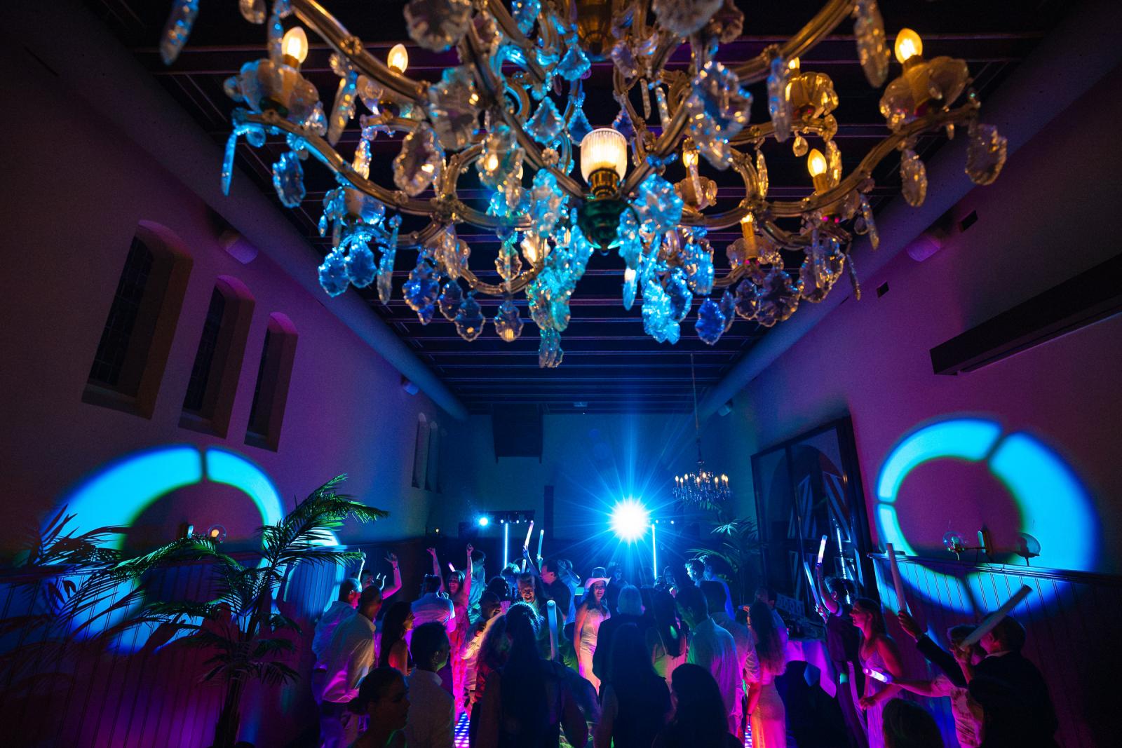 Sfeervol overzicht van de feestzaal met een grote kroonluchter op de voorgrond en een bruisende dansvloer vol gekleurd licht.