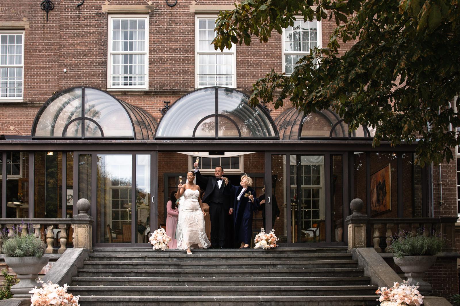Bruid en bruidegom maken een feestelijke entree op de trap bij het kasteel met glazen champagne.