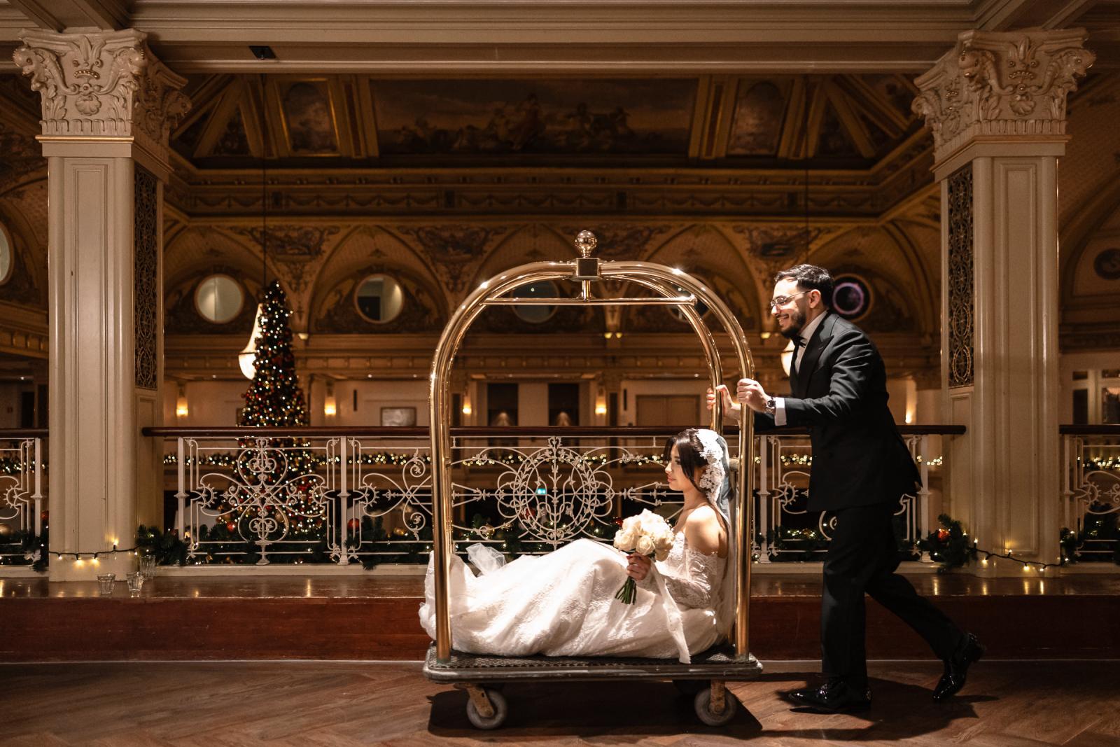 Groom playfully pushing bride in a luggage cart through the Kurhaus