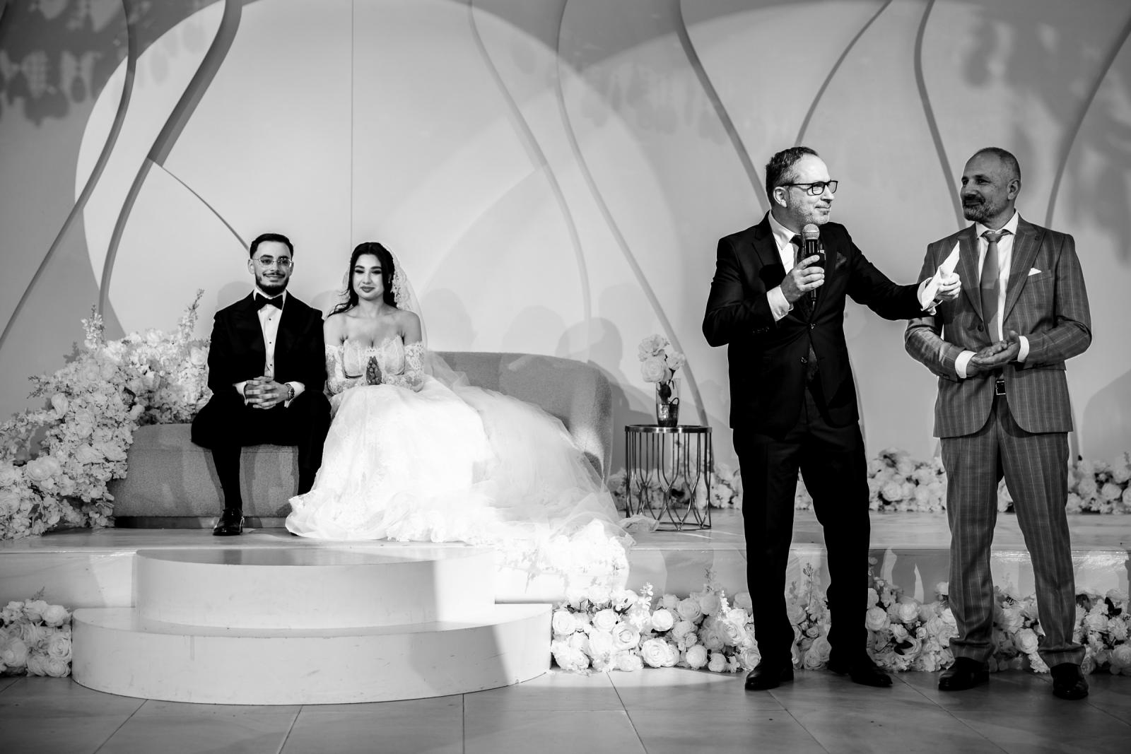The bride and groom seated on stage during speeches, with the two fathers speaking into microphones.