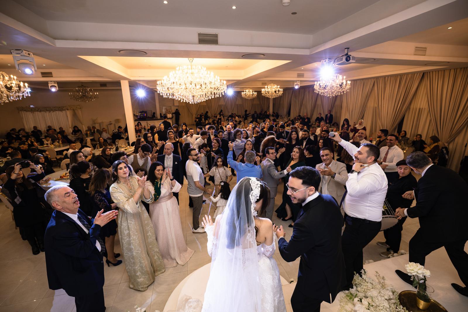 The bride and groom celebrating on stage with a large group of guests, everyone dancing and clapping.