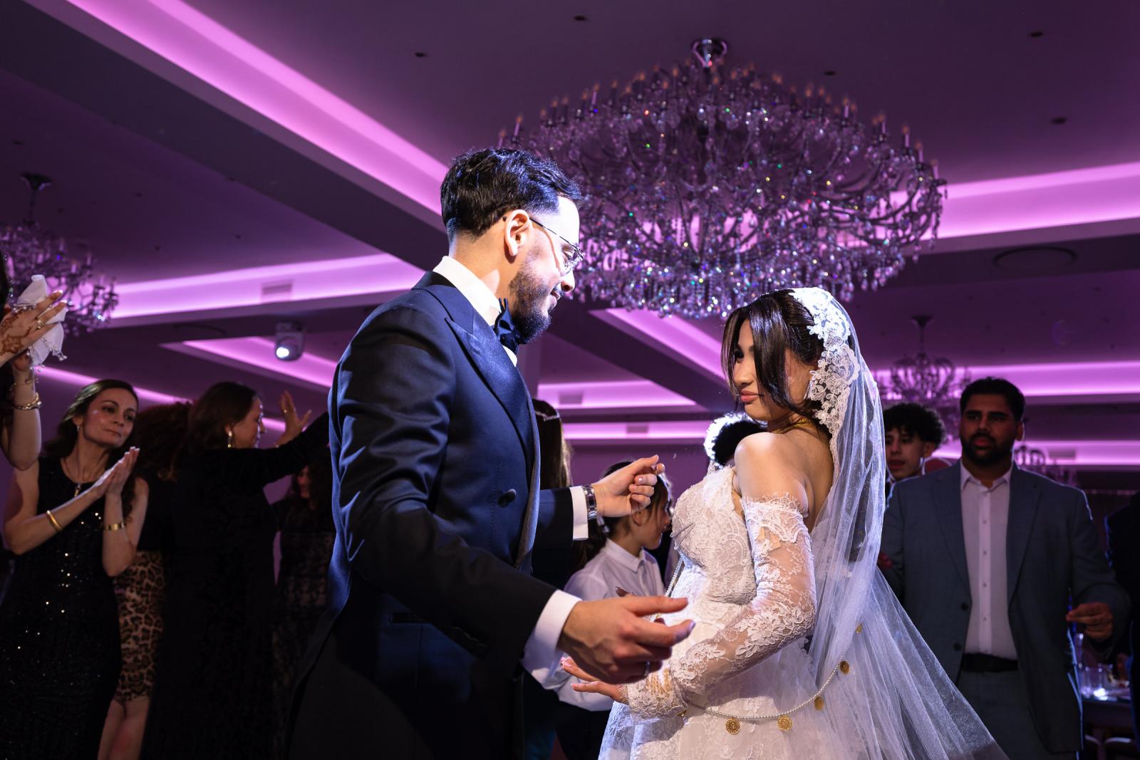 The bride and groom sharing a dance under purple lights, with guests clapping in the background.