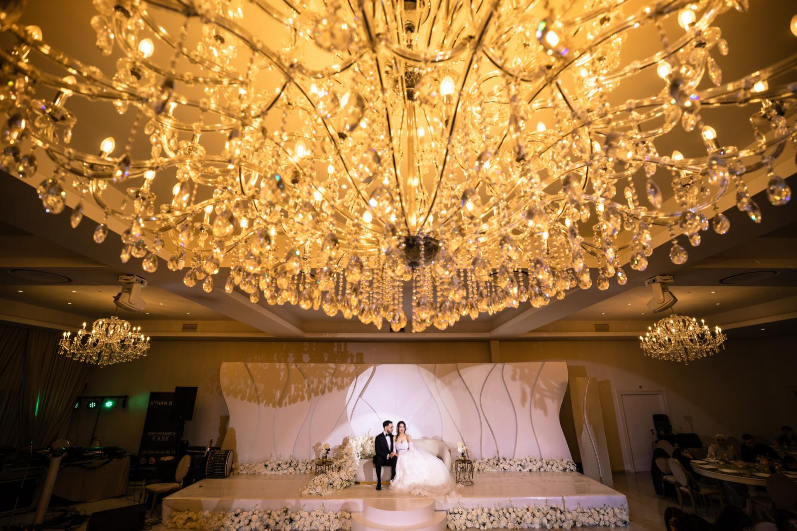 The bride and groom sitting together on a white floral stage, framed by a sparkling chandelier overhead.