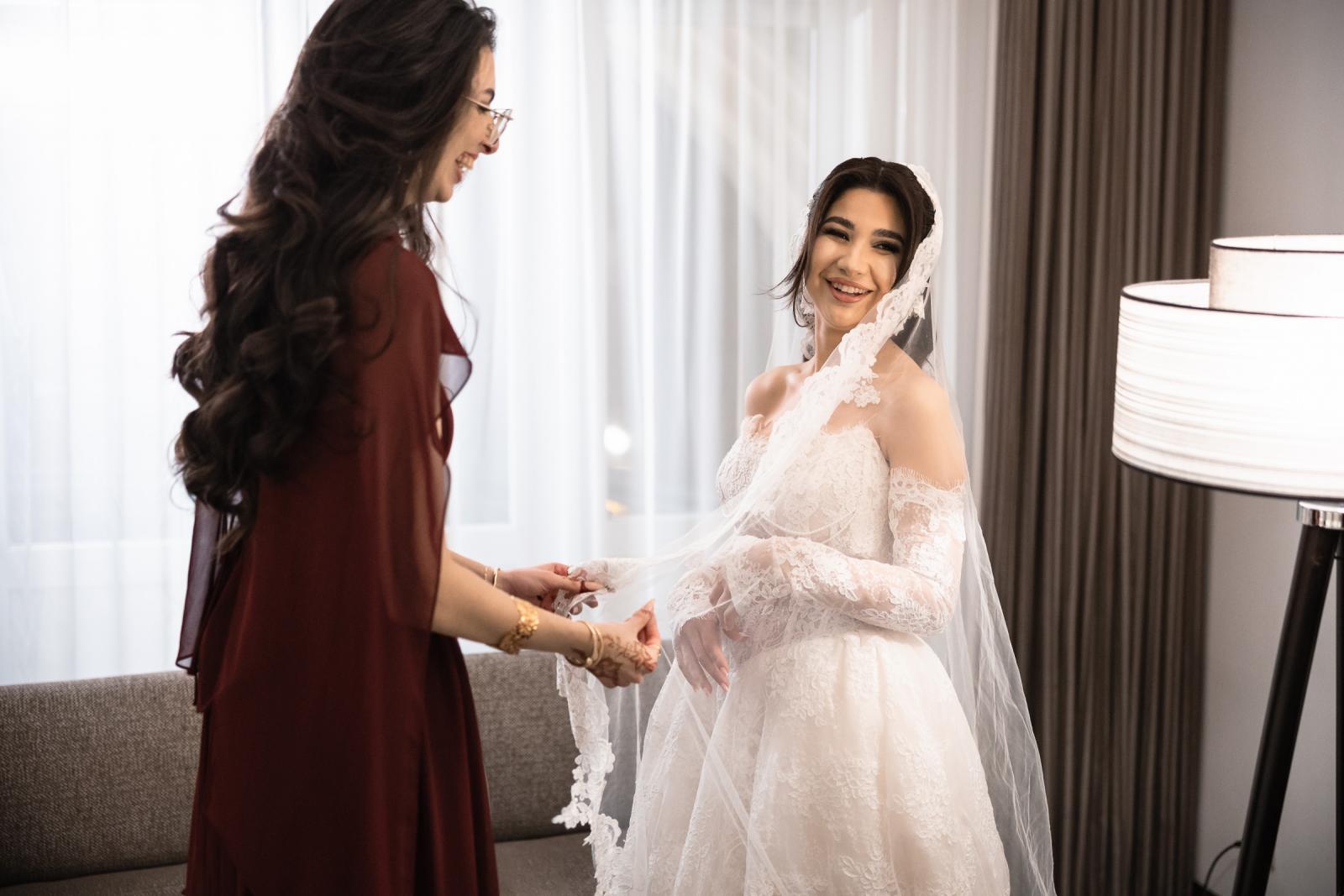 Bride smiling as her sister helps with the veil in a softly lit room