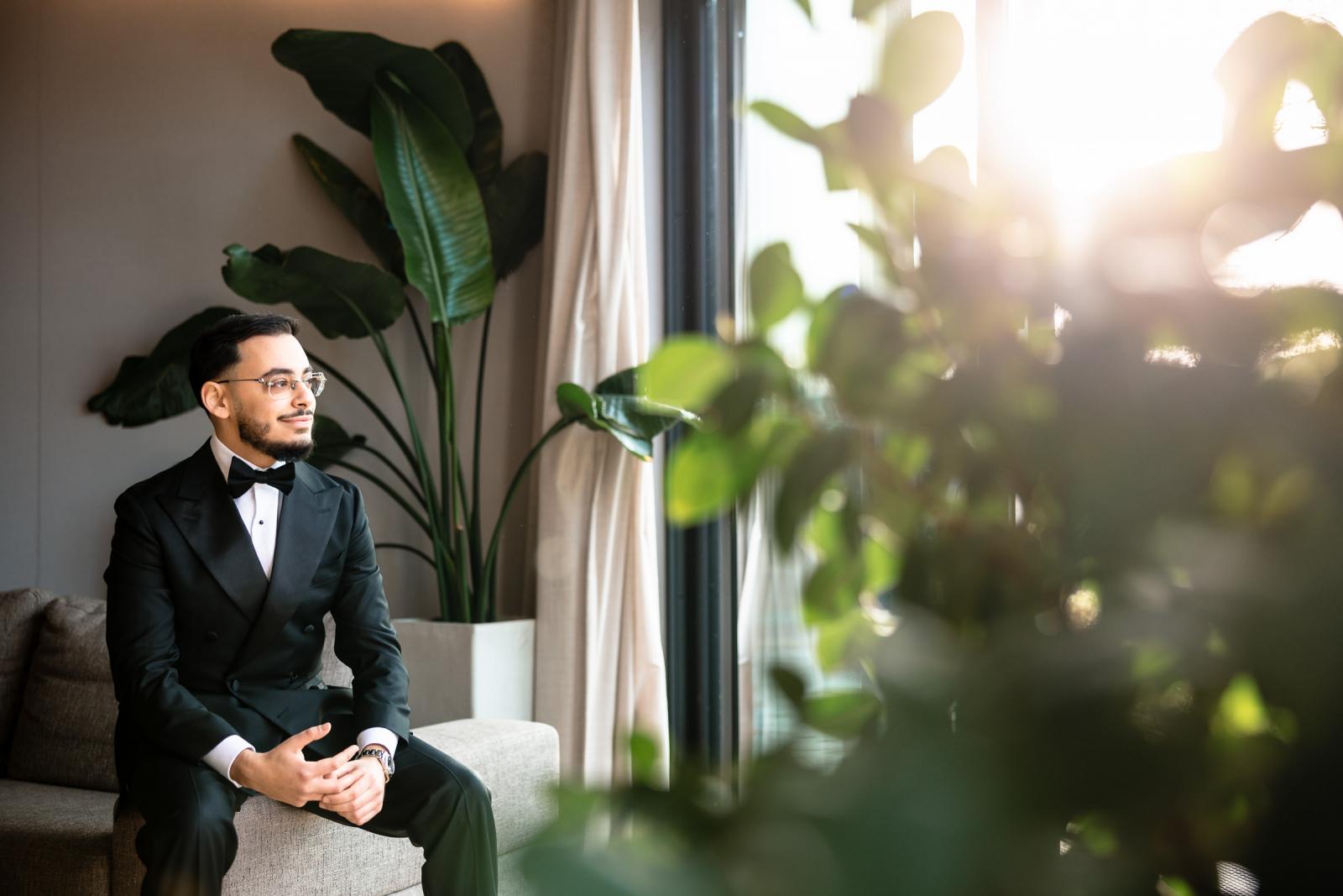 Groom sitting on the couch in a tuxedo, sunlight streaming through window and plants